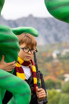 A young boy dressed as Harry Potter stands next to a giant green monster, holding a wand and looking intently at it.
