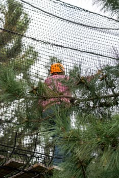 A person wearing an orange helmet and a pink sweater is climbing a rope course in a forest. They are secured by a safety harness and attached to a safety net.