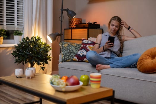 In a cozy living room, a young woman comfortably relaxes on a couch, enjoying her snacks while using her smartphone