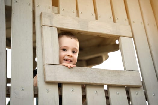 A happy smiling toddler boy of two or three years looks out of a window on a wooden playground. Toddlerhood and childhood concept. Active lifestyle