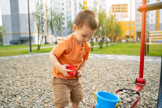 Little toddler boy playing with toys on pebbles on the playground. Boy two or three years old. Toddlerhood childhood concept. Residential complex of apartment buildings.