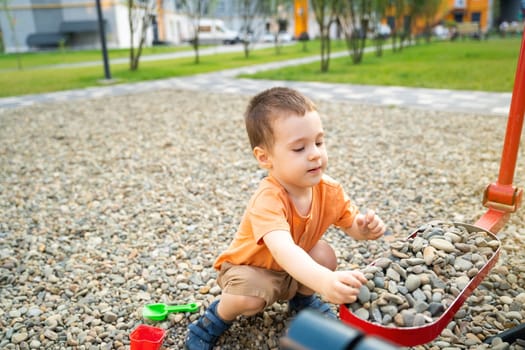 Little toddler boy playing with toys on pebbles on the playground. Boy two or three years old. Toddlerhood childhood concept. Residential complex of apartment buildings.