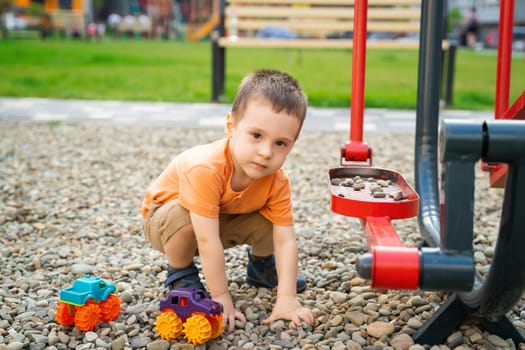 Little toddler boy playing with cars on pebbles on the playground. Boy two or three years old. Toddlerhood childhood concept. Residential complex of apartment buildings.