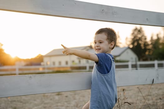 Happy toddler boy two or three years old stands near a fence and points with his finger on a sunny summer day. Summer sunset light. Horse farm