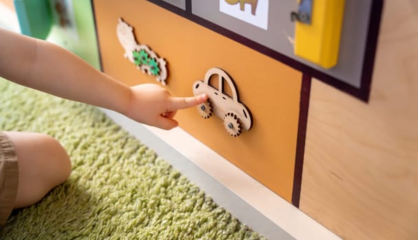 Toddler boy is playing with a busy board. Educational toy for young children. Focus on a wooden car. Close up