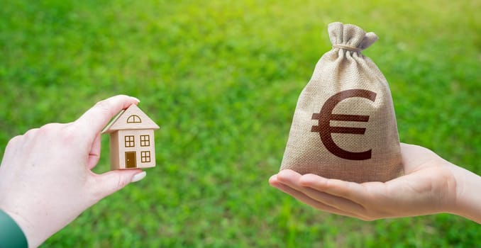 Miniature wooden house in the female hand and dollar money bag outdoors. Real estate concept. Eco-friendly home. Buying a housing outside the city.