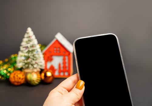 A woman's hand holds a phone with a blank screen against the background of a Christmas tree and a red house. Online shopping concept. Holiday sales and promotions. Merry christmas and happy new year