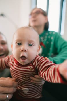 In a cozy setting, a playful baby reaches out to the camera, surrounded by loving, smiling family members