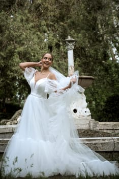 beautiful bride with long veil against the background of trees and old fountain. brunette girl in long white fluffy dress smiles and rejoices. veil flutters in the wind, bride holds veil and dress