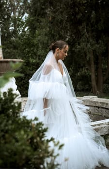 beautiful bride with long veil against the background of trees and old fountain. brunette girl in long white dress smiling. bride with smooth hair, old ruined fountain behind. girl stands sideways