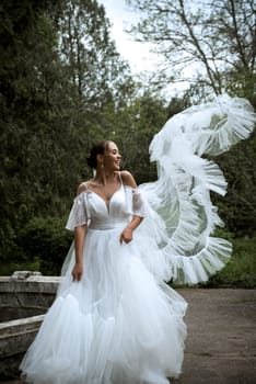 beautiful bride with long veil against the background of trees and old fountain. brunette girl in long white fluffy dress smiles and rejoices. veil flutters in the wind, full length