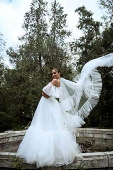beautiful bride with long veil against the background of trees and old fountain. brunette girl in long white dress rejoices. bride with smooth hair, behind old ruined fountain. veil flutters in air