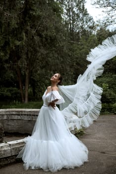 beautiful bride with long veil against the background of trees and old fountain. brunette girl in long white fluffy dress smiles and rejoices. veil flutters in the wind against