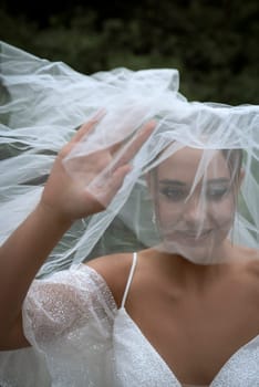 close-up of bride covered with white veil. beautiful brunette girl with sleek hairdo and stylish make-up. lifts white veil from face with hands