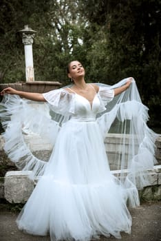 beautiful bride with long veil against the background of trees and old fountain. brunette girl in long white dress smiling. bride with smooth hair, old ruined fountain behind. girl throws veil