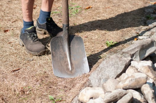 Gardener in dirty work clothes and boots holding garden tools, shovel, bunch of stones, seasonal spring garden work,