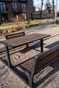 a large table and benches made of wood. benches and table are dark brown, stand in a recreation area in nature. on the ground small stones. around is clean, nearby is a pedestrian path made of tiles