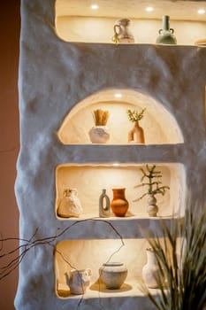 decorative wall with backlight and arches in which stand handmade vases. beige wall with shelves on them stand vases with tree branches and flowers. arches are illuminated with warm light.