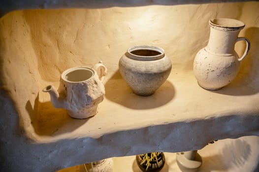 handmade clay pottery in an arch with backlight. light beige teapot, pot and jug are on the shelf. close-up of natural dishes, eco-friendly