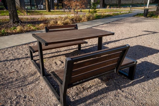 Large table and benches made of wood. Benches and table are dark brown, stand in a recreation area in nature. Small stones on the ground. It is clean around, the sun is shining. nearby pedestrian path