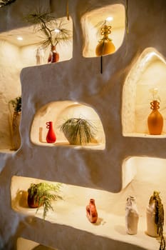 decorative wall with backlight and arches in which vases stand. beige wall in which vases with tree branches and flowers stand. arches are illuminated, handmade clay products, diagonal view