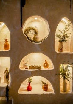 decorative wall with backlight and arches in which vases stand. beige wall in which vases with tree branches and flowers stand. arches are illuminated with warm light. handmade clay products