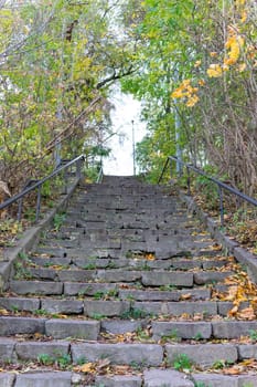 A winding staircase made of stone ascends through a natural path surrounded by trees. Colorful autumn leaves dot the ground, creating a serene atmosphere for visitors walking or hiking.