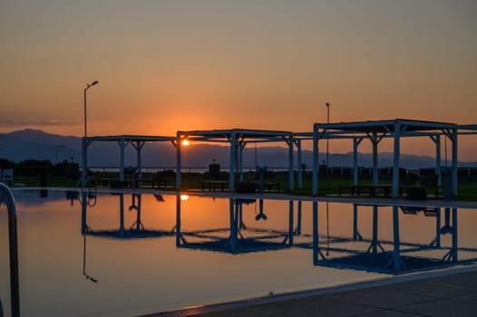 Golden sunlight dances on the calm water of a pool framed by distant mountains and tranquil structures at dusk.
