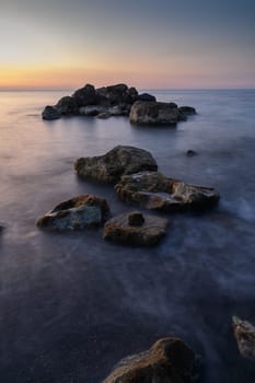 Rocks emerge from the calm water as the sun sets, casting warm hues over the tranquil shoreline at dusk.