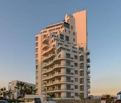 Tall, contemporary building with unique balconies captures the golden hues of sunset in a lively coastal environment.