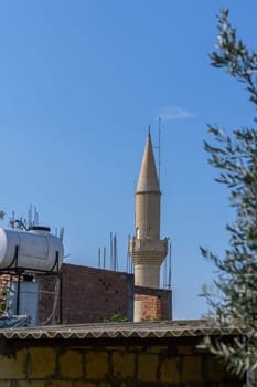 A striking minaret rises high into the blue sky, surrounded by rooftops and city structures under bright sunlight.