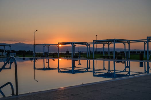 Calm waters mirror the vibrant sunset as silhouettes relax by the pool, creating a peaceful evening ambiance.