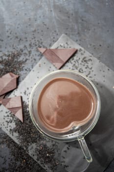 Glass cup of hot chocolate in heart shape on table surrounded by chocolate pieces and black chia seeds on grey textured background, Desert concept in color of the year 2025, mocha mousse, copy space