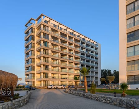 Sunset casts a warm glow over a sleek residential building set amidst lush greenery and palm trees by the coast.