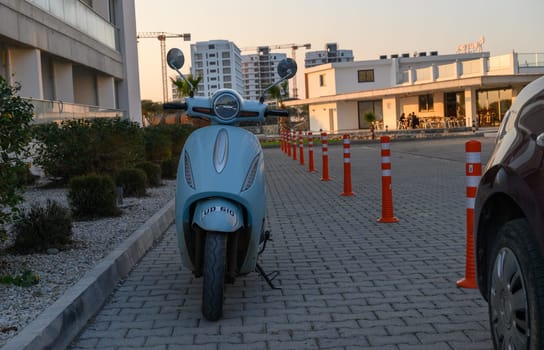 A bright blue scooter stands parked beside a paved path as the sun sets behind contemporary buildings.