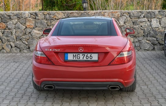 Gaziveren Cyprus -30.01.2025 Bright red convertible stands proudly in front of a textured stone wall surrounded by greenery at sunset.