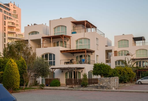 A stunning modern building with large windows glows warmly as the sun sets, surrounded by lush gardens and trees.