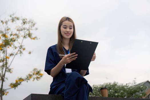 A happy nurse in scrubs sits outdoors, smiling while reviewing patient notes on a clipboard, symbolizing dedication to healthcare and documentation.