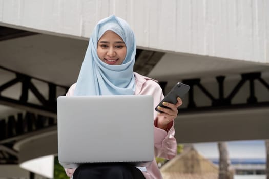 A focused Muslim woman in a hijab smiles while using her laptop and smartphone, representing modern business and technology integration.