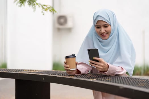 A joyful Muslim woman in a hijab holds a coffee cup and checks her smartphone, representing a balanced lifestyle and modern business engagement.