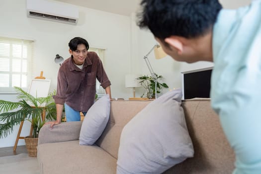 Two men joyfully arranging their new living room, one leaning on the sofa with a smile, surrounded by plants and a bright, welcoming atmosphere.