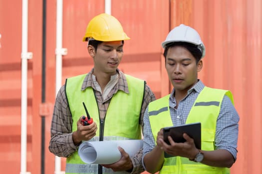 Two professionals in safety vests discuss project plans, one holding blueprints and the other a tablet, highlighting collaboration in logistics and engineering.