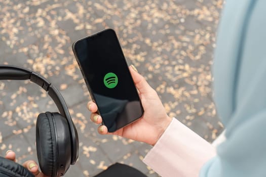 A person using a smartphone featuring the Spotify logo while holding headphones outdoors amidst fallen leaves, enjoying music.