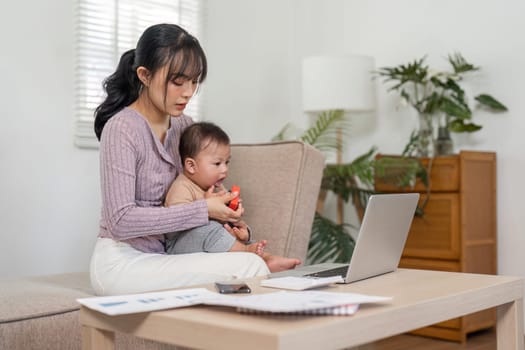 A mother balancing work on her laptop while holding her baby and managing paperwork in a cozy home setting, showcasing modern parenting.
