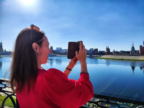 A woman in a red dress captures a beautiful skyline along the water under clear blue skies.