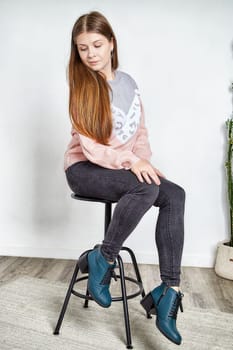 Young model poses gracefully on a stool, highlighting trendy shoes and fashionable attire in a contemporary space.