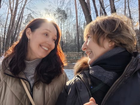 Two friends share a joyful moment outdoors, smiling at each other in a bright, sunny winter landscape surrounded by trees.