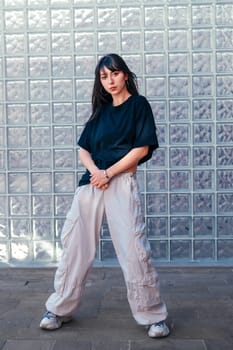 A young woman poses confidently in casual attire, featuring a dark shirt and light cargo pants, in front of a textured glass wall in an urban environment, showcasing her unique style.