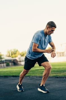 A man in a blue shirt and shorts checks his smartwatch while jogging on a track field. The scene is set during the afternoon with clear skies and green grass in the background.