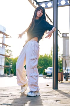 A young woman showcases her dance moves in an urban environment as the sun sets behind her. She wears loose, casual clothing and sneakers, exuding a vibrant energy.